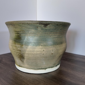 Pode incluir: Um vaso de cerâmica artesanal com uma borda larga e ligeiramente alargada. O exterior apresenta uma mistura de tons verdes e bege, com uma base branca. A cerâmica tem uma superfície texturizada, mostrando as variações naturais da argila.
