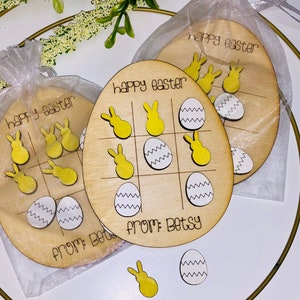 May include: Two wooden Easter tic-tac-toe game boards with yellow bunny and white egg pieces. The boards are oval shaped and have the text "happy easter" and "from: Betsy" on them.