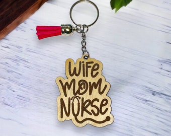 May include: A wooden keychain with the words "Wife Mom Nurse" engraved in a cursive font. The keychain has a silver ring and chain, and a bright pink tassel. The wood has a natural, light brown color.