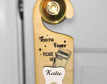 May include: A wooden tooth fairy door hanger with a gold bell, a slot for a tooth, and the text "Tooth Fairy Please Leave Her" and a tooth with the name "Katie" on it.