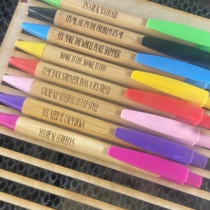 K&ouml;nnte beinhalten: Ein Set aus zehn Bambus-Stiften mit bunten Kappen und inspirierenden Zitaten, die in den Schaft graviert sind. Die Zitate beinhalten "Ich bin ein echter harter Kerl", "Du l&auml;sst den ganzen Ort gl&auml;nzen", "Komm st&auml;rker zur&uuml;ck als ein 90er-Jahre-Trend" und "Du bist so wundersch&ouml;n".