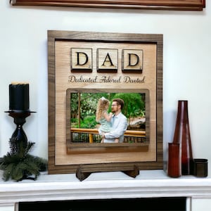 May include: A wooden picture frame with a photo of a father and daughter. The frame has a scrabble tile inscription that reads "DAD" and the words "Dedicated, Adored, Devoted".