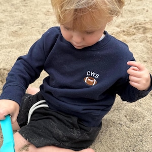 May include: A navy blue long-sleeve sweatshirt featuring an embroidered brown football graphic with the white letters CWB positioned above it. The sweatshirt has a crew neckline and ribbed cuffs, providing a simple and functional design.