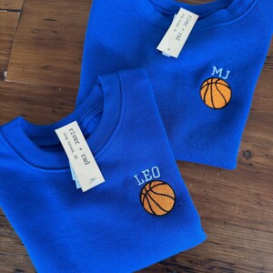 May include: Two royal blue crewneck sweatshirts featuring embroidered basketball designs. One sweatshirt is personalised with the name LEO and the other with the initials MJ in light blue thread. Each garment includes a white river + rad brand tag.