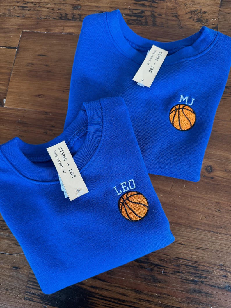 May include: Two royal blue crewneck sweatshirts featuring embroidered basketball designs. One sweatshirt is personalised with the name LEO and the other with the initials MJ in light blue thread. Each garment includes a white river + rad brand tag.