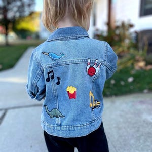 May include: A blue denim jacket with various embroidered patches, including a narwhal, musical notes, french fries, a bowling pin, a dinosaur, and a construction vehicle.
