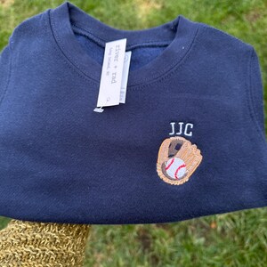 May include: A navy blue crewneck sweatshirt featuring an embroidered design of a tan baseball glove holding a white baseball with red stitching. Above the glove, the letters JJC are embroidered in white. A white brand tag reads river + rad.
