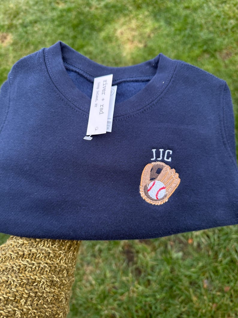 May include: A navy blue crewneck sweatshirt featuring an embroidered design of a tan baseball glove holding a white baseball with red stitching. Above the glove, the letters JJC are embroidered in white. A white brand tag reads river + rad.