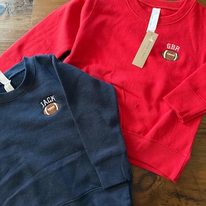May include: A red long-sleeve sweatshirt with an embroidered brown football and the text GBR, and a navy blue long-sleeve sweatshirt with an embroidered brown football and the text JACK. Both garments feature crew necklines and ribbed cuffs.