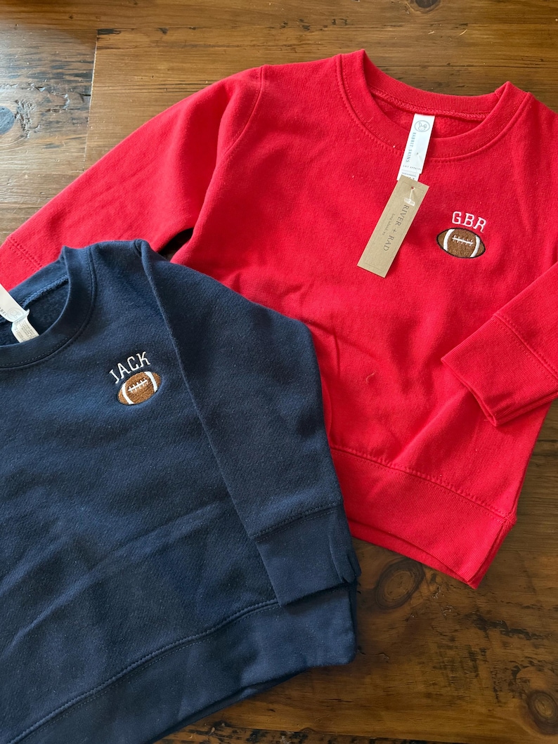 May include: A red long-sleeve sweatshirt with an embroidered brown football and the text GBR, and a navy blue long-sleeve sweatshirt with an embroidered brown football and the text JACK. Both garments feature crew necklines and ribbed cuffs.