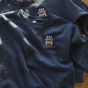 May include: Two navy blue crewneck sweatshirts featuring an embroidered design on the upper left chest. The embroidery consists of the white text "WAR EAGLE" arched over a brown and white football graphic with white stitching details.
