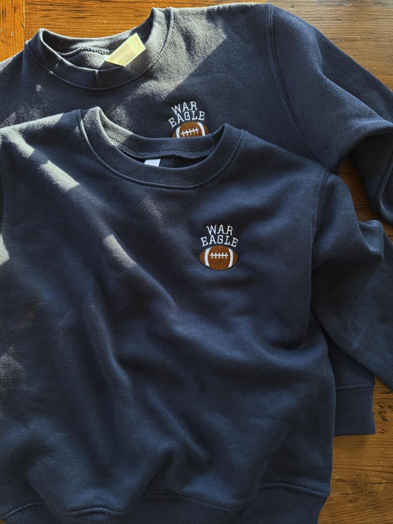 May include: Two navy blue crewneck sweatshirts featuring an embroidered design on the upper left chest. The embroidery consists of the white text "WAR EAGLE" arched over a brown and white football graphic with white stitching details.