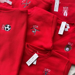 May include: A collection of bright red crewneck sweatshirts, each featuring unique embroidered designs including a football, ballet slippers, a hockey stick, a gymnast, a baseball glove, and skis, with white tags labelled river + rad attached.