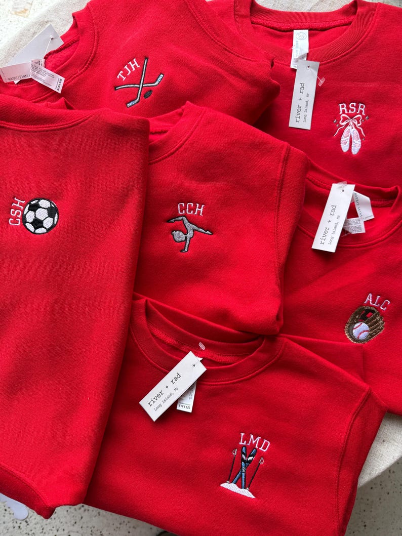 May include: A collection of bright red crewneck sweatshirts, each featuring unique embroidered designs including a football, ballet slippers, a hockey stick, a gymnast, a baseball glove, and skis, with white tags labelled river + rad attached.