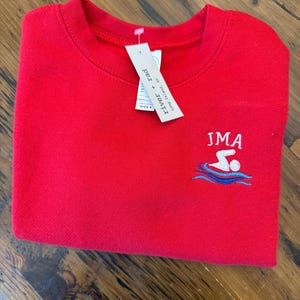 May include: A bright red crewneck sweatshirt featuring an embroidered graphic of a person swimming in blue waves with the text JMA above it. A white retail tag attached to the collar displays the brand name river + rad and the size 4.