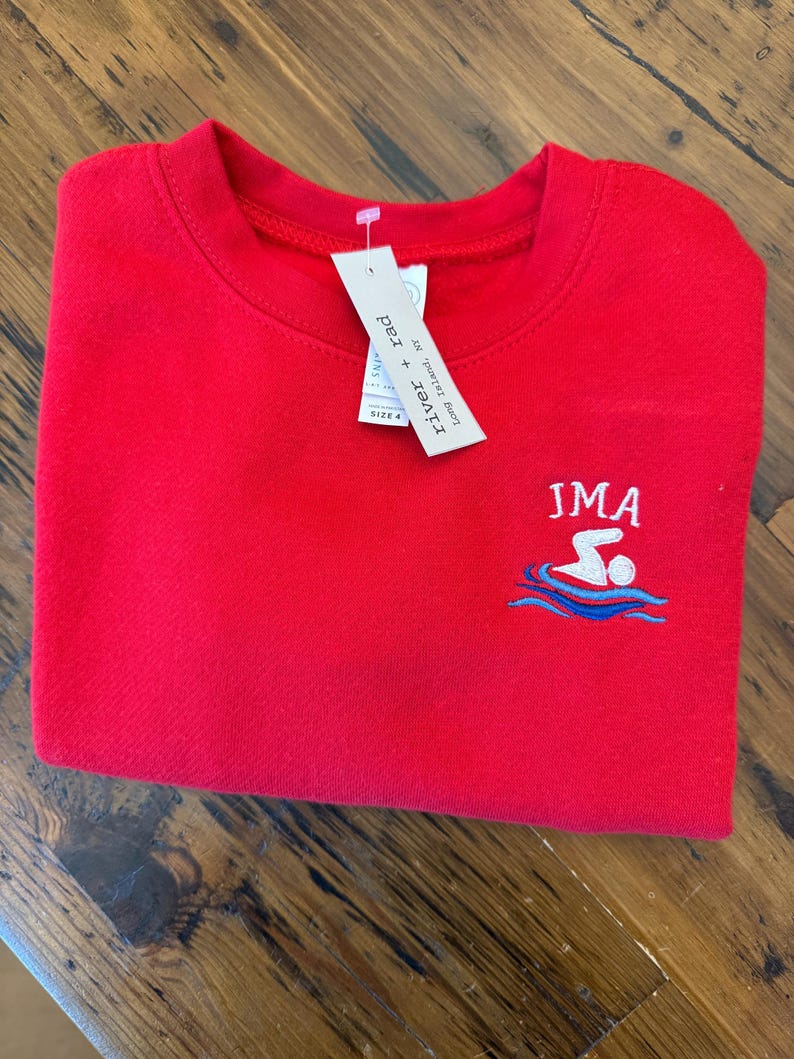 May include: A bright red crewneck sweatshirt featuring an embroidered graphic of a person swimming in blue waves with the text JMA above it. A white retail tag attached to the collar displays the brand name river + rad and the size 4.