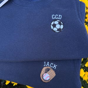 May include: Two navy blue sweatshirts featuring embroidered designs. The top sweatshirt displays a football with the letters CCD above it. The bottom sweatshirt features a baseball glove holding a baseball with the name JACK embroidered above it.