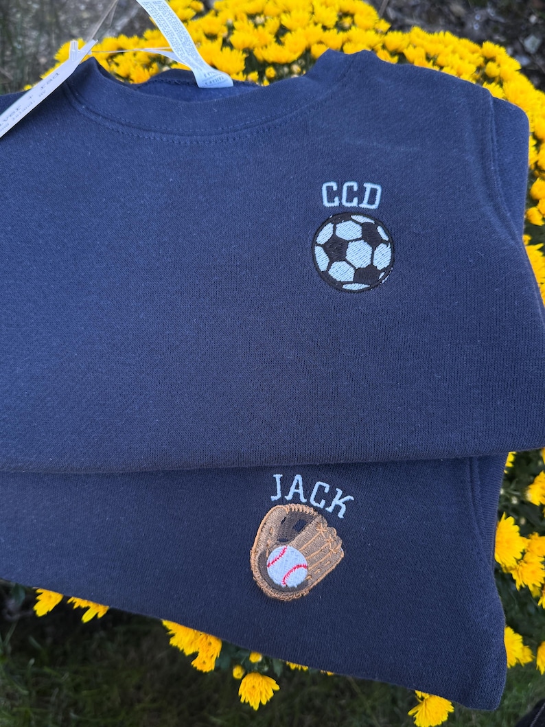 May include: Two navy blue sweatshirts featuring embroidered designs. The top sweatshirt displays a football with the letters CCD above it. The bottom sweatshirt features a baseball glove holding a baseball with the name JACK embroidered above it.