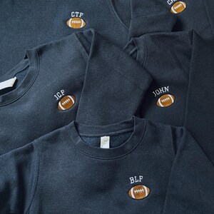 May include: A collection of dark charcoal grey crewneck sweatshirts, each featuring an embroidered brown football graphic on the chest. Above each football, white embroidered text displays the initials CTF, CHF, JCP, JOHN, or BLF.