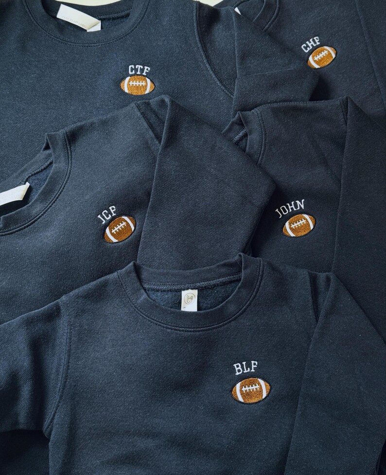 May include: A collection of dark charcoal grey crewneck sweatshirts, each featuring an embroidered brown football graphic on the chest. Above each football, white embroidered text displays the initials CTF, CHF, JCP, JOHN, or BLF.