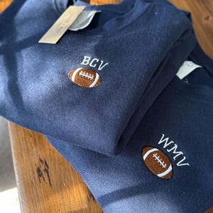 May include: Two navy blue crewneck sweatshirts featuring embroidered designs on the chest. Each sweatshirt displays a brown football icon with white stitching details, accompanied by white embroidered letters reading BCV on one and WMV on the other.