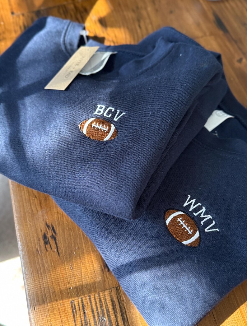 May include: Two navy blue crewneck sweatshirts featuring embroidered designs on the chest. Each sweatshirt displays a brown football icon with white stitching details, accompanied by white embroidered letters reading BCV on one and WMV on the other.