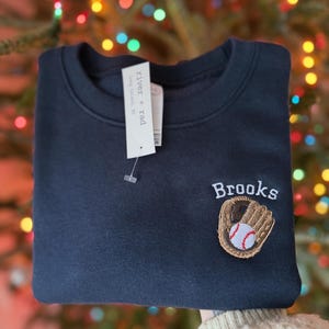 May include: A dark navy blue crewneck sweatshirt featuring an embroidered brown baseball glove holding a white baseball with red stitching. Above the glove, the name Brooks is embroidered in white thread. A white tag reads river + rad.