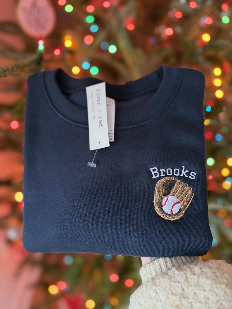 May include: A dark navy blue crewneck sweatshirt featuring an embroidered brown baseball glove holding a white baseball with red stitching. Above the glove, the name Brooks is embroidered in white thread. A white tag reads river + rad.