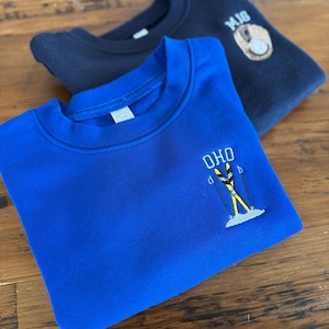 May include: A bright blue crewneck sweatshirt featuring an embroidered design of yellow skis, poles, and the text OHO, layered over a dark navy sweatshirt with an embroidered baseball glove, ball, and the letters MJG on the upper chest area.