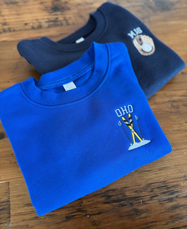 May include: A bright blue crewneck sweatshirt featuring an embroidered design of yellow skis, poles, and the text OHO, layered over a dark navy sweatshirt with an embroidered baseball glove, ball, and the letters MJG on the upper chest area.