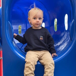 May include: A navy blue long-sleeve sweatshirt featuring the letters CWB and a small brown football graphic embroidered on the left chest. The sweatshirt is paired with tan-coloured trousers with elastic cuffs and black and white slip-on shoes.