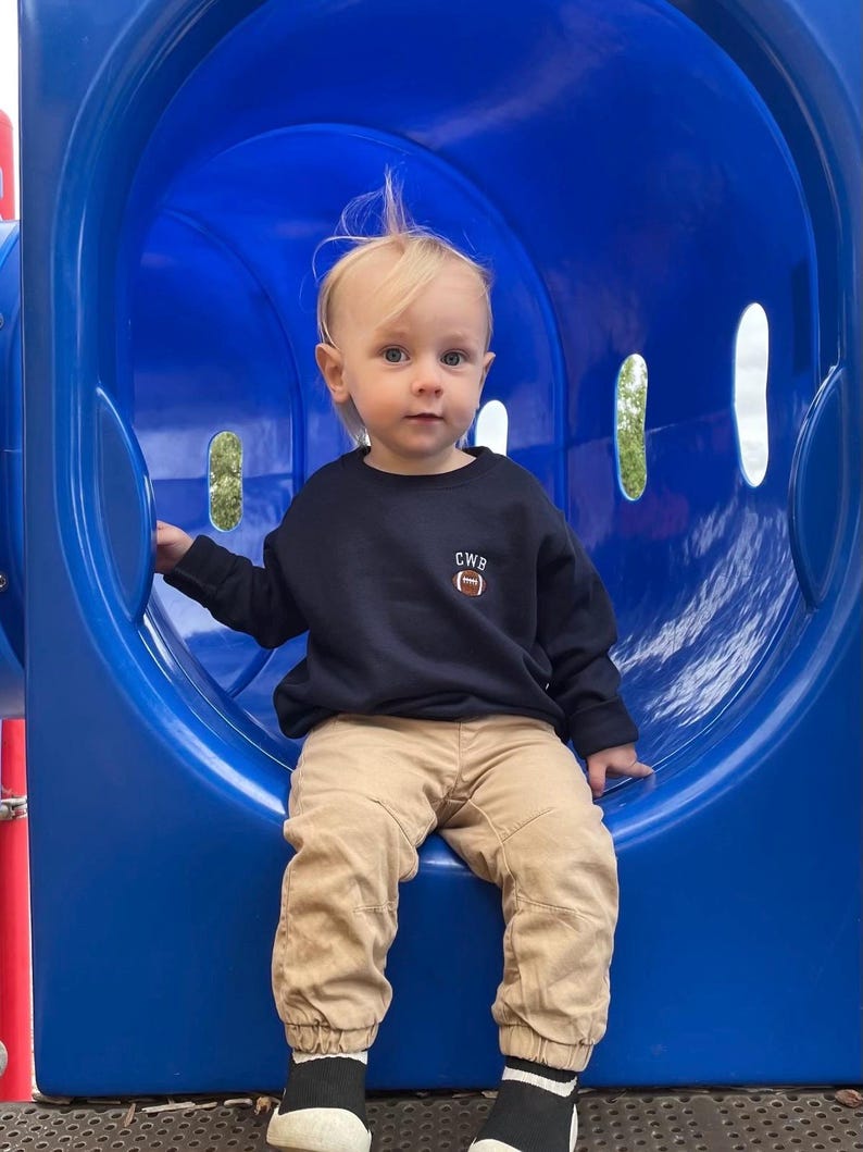 May include: A navy blue long-sleeve sweatshirt featuring the letters CWB and a small brown football graphic embroidered on the left chest. The sweatshirt is paired with tan-coloured trousers with elastic cuffs and black and white slip-on shoes.