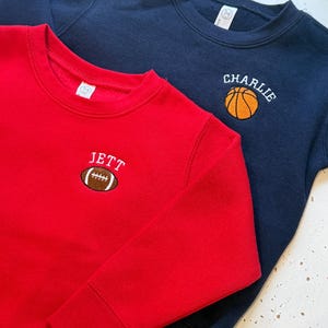 May include: A red sweatshirt featuring an embroidered brown football with the name JETT above it, and a navy blue sweatshirt featuring an embroidered orange basketball with the name CHARLIE above it. Both garments have long sleeves and crew necklines.