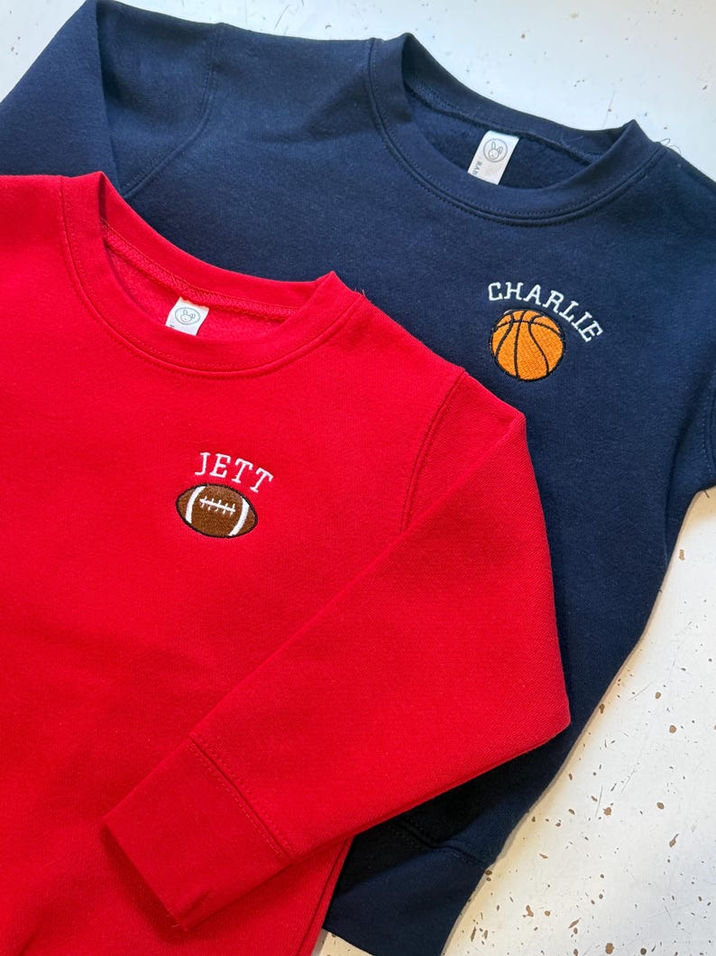 May include: A red sweatshirt featuring an embroidered brown football with the name JETT above it, and a navy blue sweatshirt featuring an embroidered orange basketball with the name CHARLIE above it. Both garments have long sleeves and crew necklines.