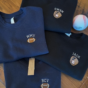 May include: Four crewneck sweatshirts in navy blue and black, each featuring custom embroidery. Two feature a brown football with initials WMV or BCV, and two feature a baseball glove with a ball and the names OWEN or JACK. A baseball is included.