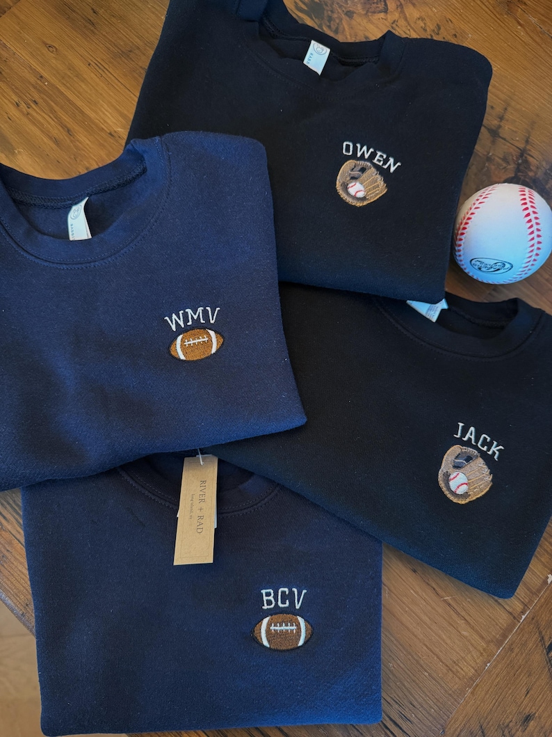May include: Four crewneck sweatshirts in navy blue and black, each featuring custom embroidery. Two feature a brown football with initials WMV or BCV, and two feature a baseball glove with a ball and the names OWEN or JACK. A baseball is included.