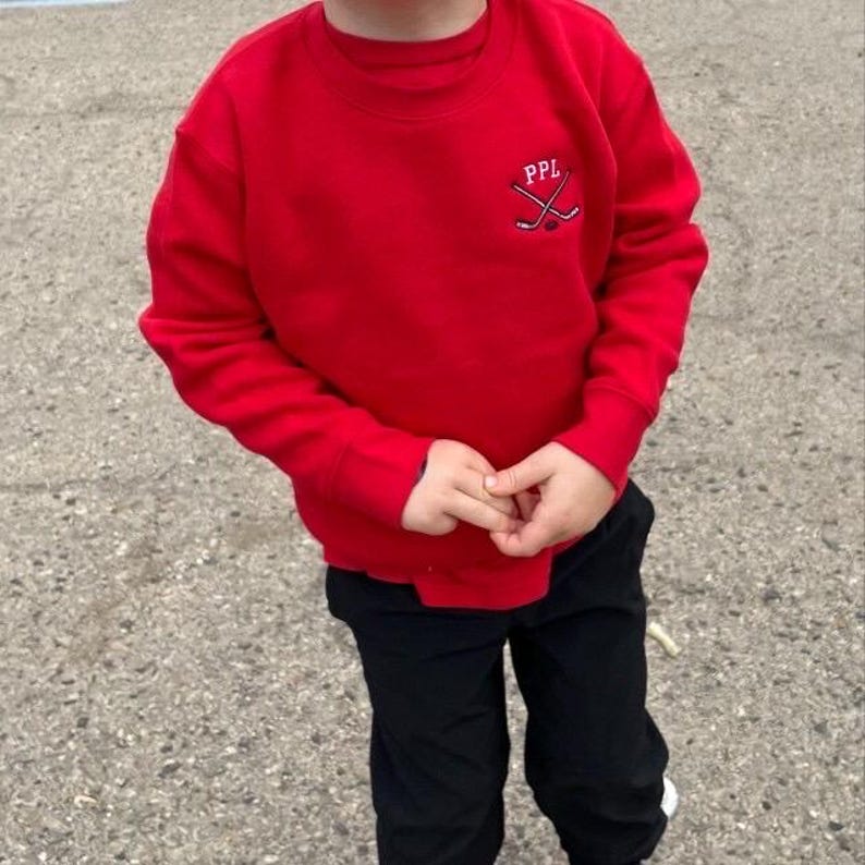 May include: A bright red long-sleeve crewneck sweatshirt featuring an embroidered logo on the upper left chest. The logo consists of the white text PPL positioned above two crossed white hockey sticks and a small black hockey puck graphic.