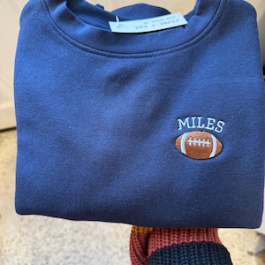 May include: A navy blue crewneck sweatshirt featuring an embroidered design on the upper right chest. The embroidery consists of a brown football with white laces and the name MILES stitched in white capital letters above the football.