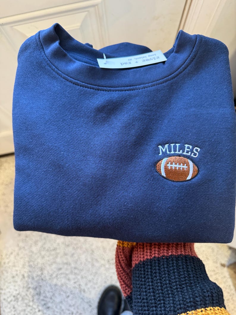 May include: A navy blue crewneck sweatshirt featuring an embroidered design on the upper right chest. The embroidery consists of a brown football with white laces and the name MILES stitched in white capital letters above the football.