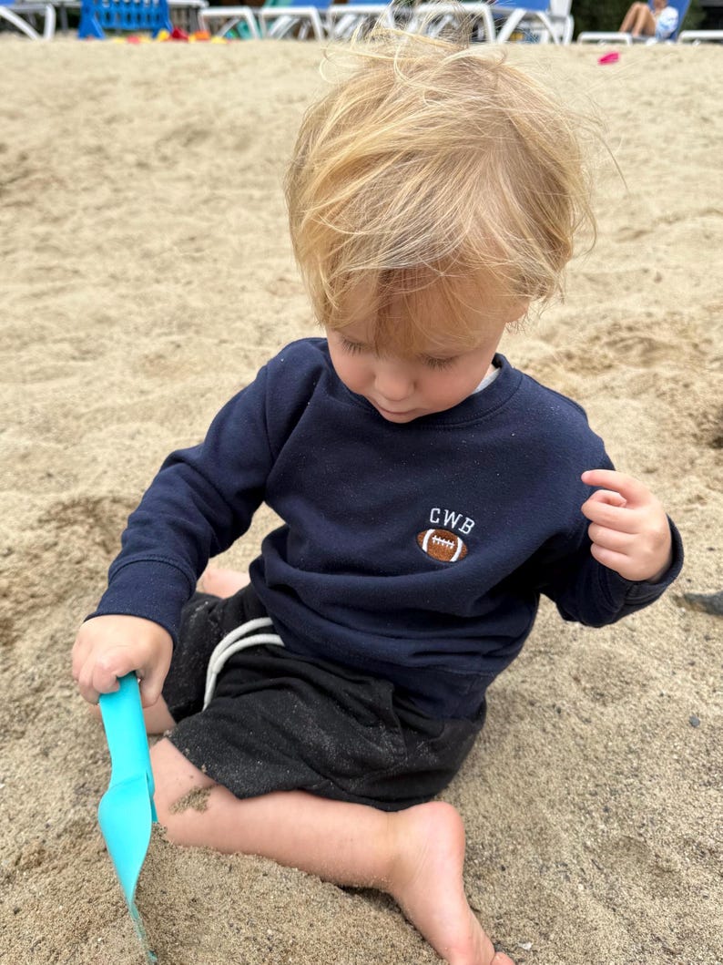 May include: A navy blue long-sleeve sweatshirt featuring an embroidered brown football graphic with the white letters CWB positioned above it. The sweatshirt has a crew neckline and ribbed cuffs, providing a simple and functional design.