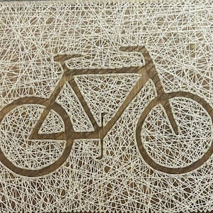 May include: A white string art piece depicting a bicycle on a brown wooden background.