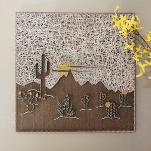 May include: A string art piece depicting a desert landscape with a setting sun, mountains, and cacti. The art is made with white string wrapped around nails hammered into a brown fabric background.