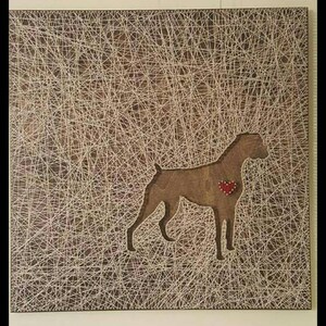 May include: A string art piece depicting a brown dog with a red heart on its chest. The dog is outlined with white string against a brown wooden background.