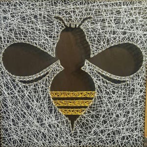 May include: A white string art design of a bee on a dark brown background. The bee has a yellow and gold striped abdomen.