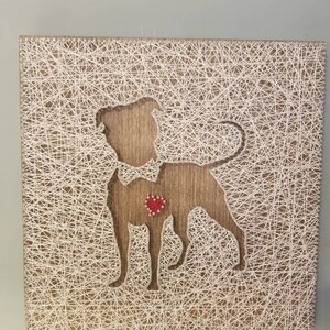 May include: A string art piece depicting a dog with a red heart in the center. The dog is outlined in white string on a brown wooden background.