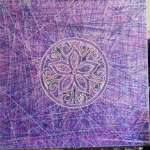 May include: A purple and white string art piece with a circular design featuring a flower in the center. The background is a criss-cross pattern of string.