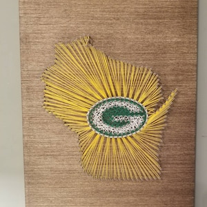 May include: A string art piece depicting the state of Wisconsin with a green and white "G" in the center. The state outline is made of yellow string, and the "G" is made of green, white, and gray string.