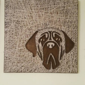 May include: A brown wooden frame with white string art depicting the head of a dog. The dog's face is outlined in brown wood.