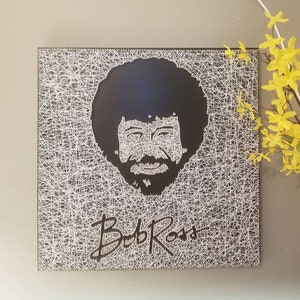 May include: A black and white string art portrait of Bob Ross, featuring his signature afro hairstyle and a smile. The artwork is framed and includes his signature "Bob Ross" in white string.