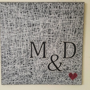 May include: A black canvas with white string art forming a design of the letters M and D with an ampersand between them. A small red heart is at the bottom of the design.